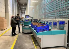 Espace produits secs comportant huit postes de préparation. L'opératrice récupère les produits acheminés par les robots pour les placer dans les bacs bleus. 